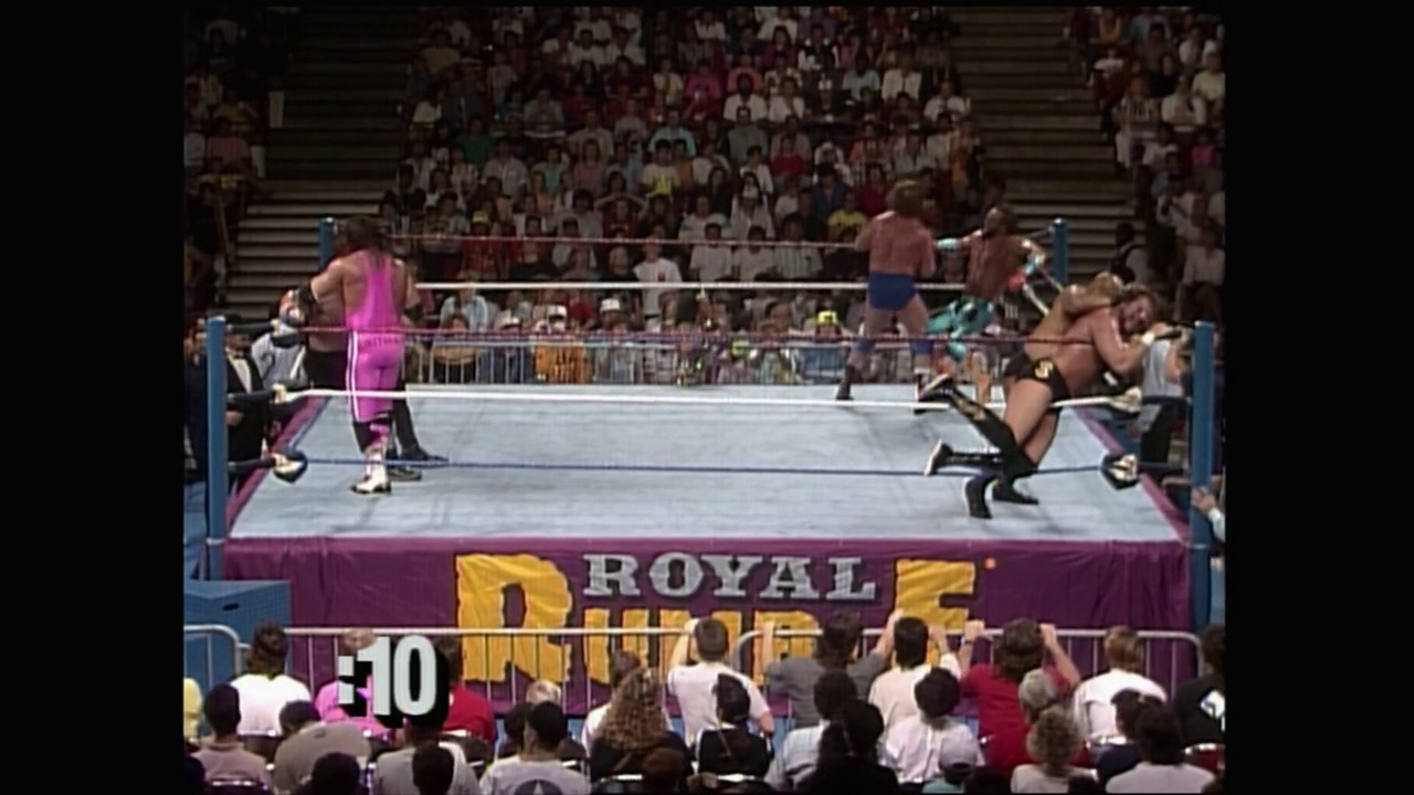 A frame from Royal Rumble (1990) at 1:55:31. A professional wrestling match is underway in a WWF Royal Rumble ring, with multiple shirtless wrestlers in the squared circle engaging in combat while a packed stadium crowd watches from the stands above. The burgundy ring apron displays the "Royal Rumble" branding in yellow lettering.