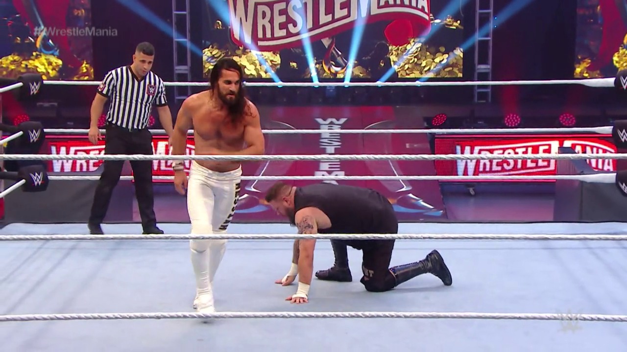A frame from WrestleMania 36 (2020) at 1:56:29. # WrestleMania 36 Ring Action

A wrestler in white pants stands in the WWE ring while an opponent in black crawls on the mat, with a referee in striped shirt overseeing the match. The vibrant WrestleMania branding and purple lighting illuminate the arena backdrop.