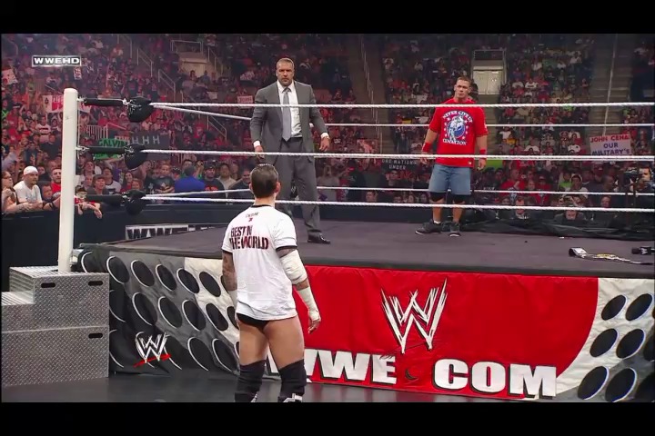 A frame from WWE Raw (August 8, 2011) at 1:36:47. A WWE wrestler in a white shirt stands outside the ring facing two opponents inside, with a full crowd visible in the background and the WWE logo displayed on the ring apron.