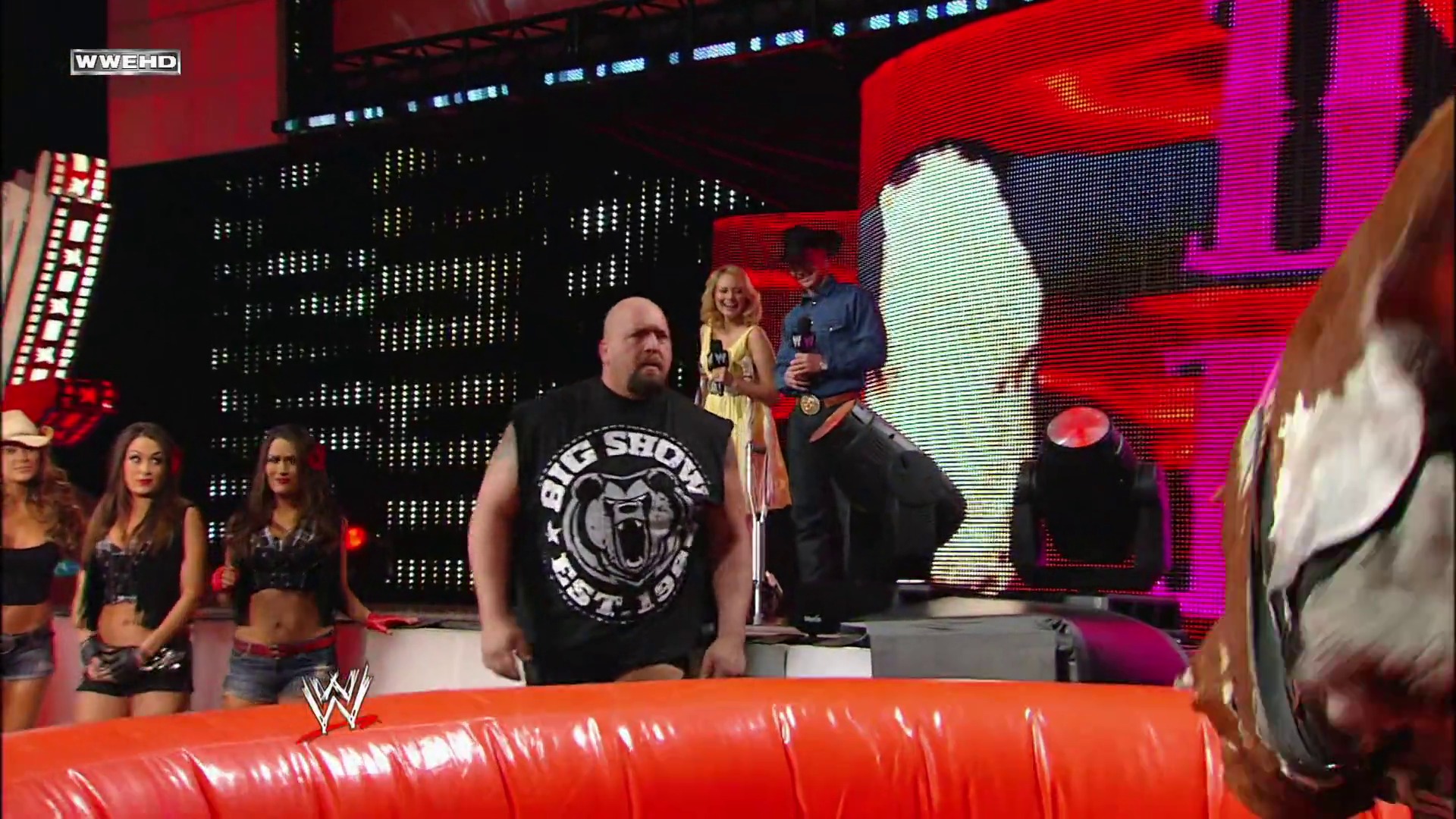 A frame from WWE Raw (February 22, 2010) at 1:07:56. A WWE Raw event scene featuring a bald wrestler in a black "Big Show" t-shirt standing in the ring, with female performers in the background, elaborate LED screens displaying graphics, and an energetic arena setting with red and pink lighting.