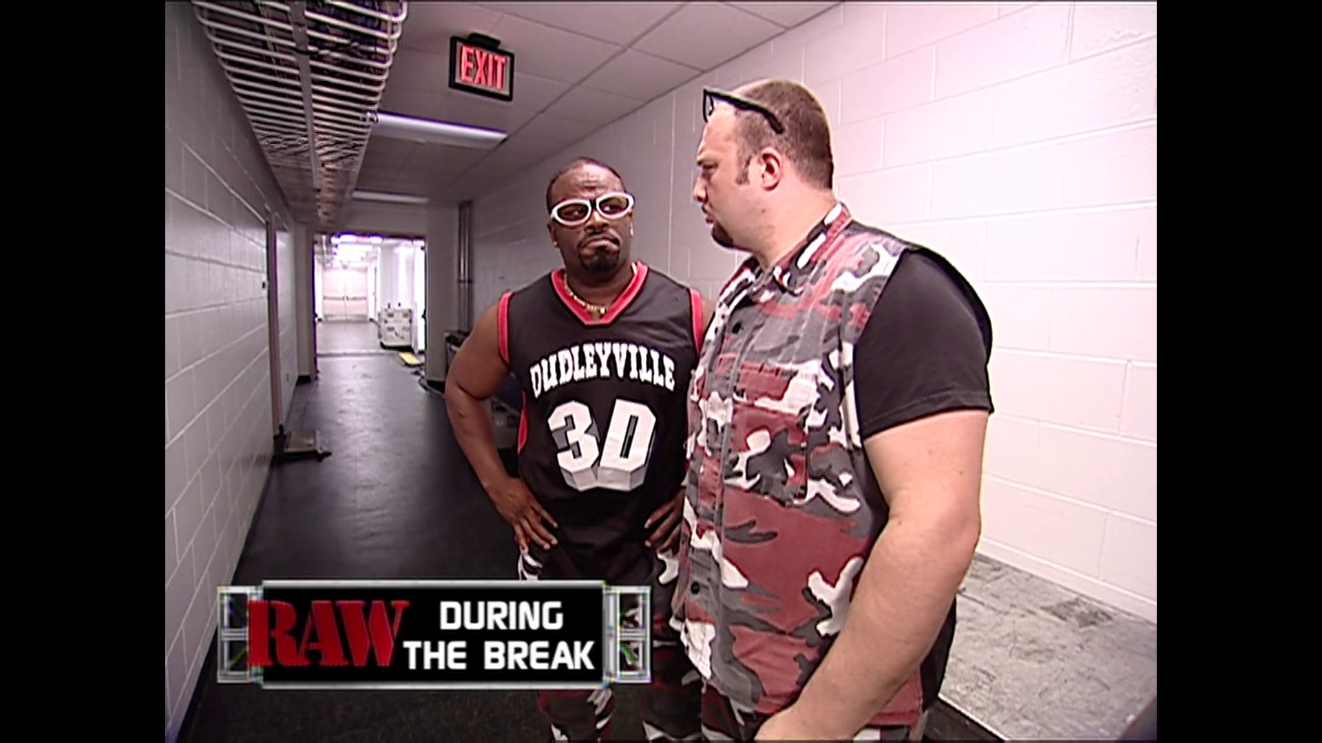 A frame from WWE Raw (June 18, 2001) at 14:40. Two men converse in a backstage hallway during WWE Raw. One wears a "Dudleyville 30" basketball jersey and sunglasses, while the other sports a patterned vest over a black shirt, with a "RAW DURING THE BREAK" caption visible at the bottom.