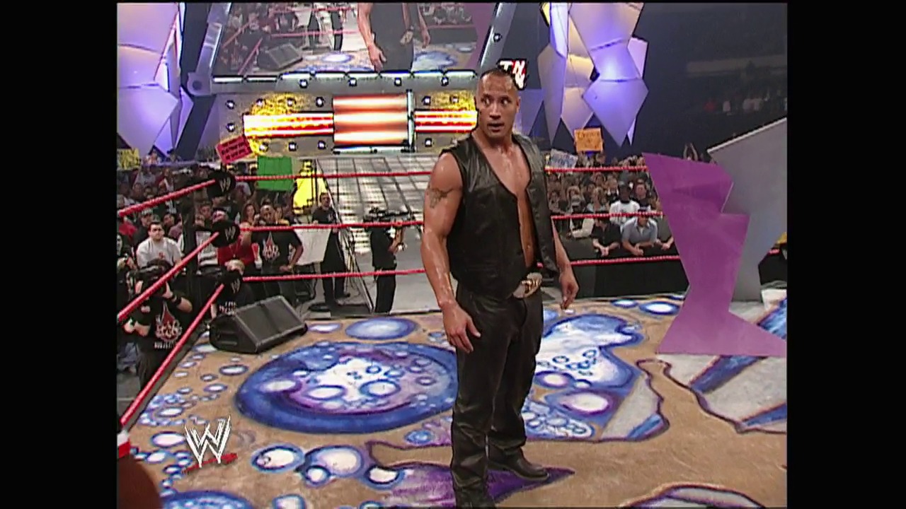 A frame from WWE Raw (June 2, 2003) at 53:07. A muscular wrestler in dark attire stands in the WWE Raw ring surrounded by blue and purple circular graphics on the mat. The packed arena with red ropes is visible in the background, with the WWE logo displayed in the lower left corner.