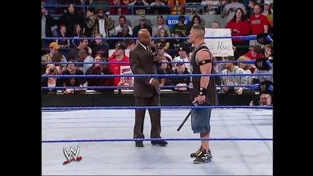 A frame from WWE SmackDown (March 11, 2005) at 35:36. Two wrestlers stand in a WWE ring during SmackDown, with one in a brown suit and the other in dark attire holding a weapon. A packed crowd fills the arena stands in the background.