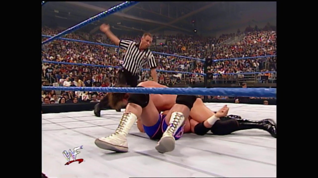 A frame from WWE SmackDown (May 25, 2000) at 35:31. A wrestler lies on the mat wearing white boots and blue/purple wrestling attire while a referee in black and white stripes stands over them with his arm raised. A packed stadium crowd fills the background of the blue-roped WWE ring.
