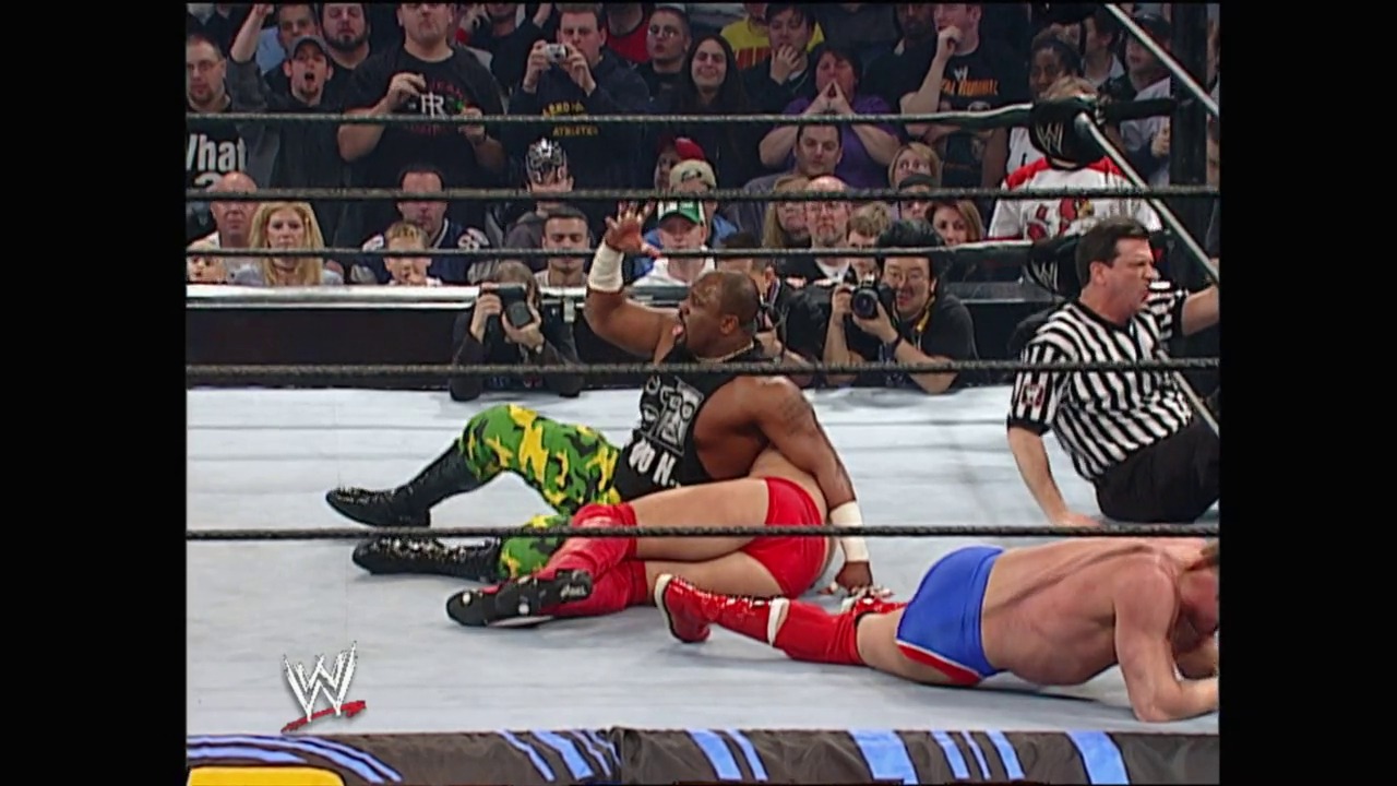 A frame from Royal Rumble (2003) at 23:14. Two wrestlers lie on the mat during a match at WWE Royal Rumble 2003, with a referee in striped shirt nearby and an engaged crowd visible behind the ring ropes.