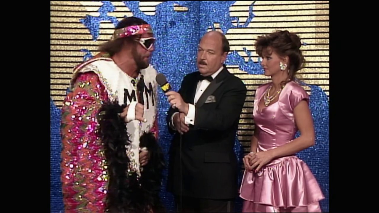 A frame from WrestleMania IV (1988) at 1:59:48. # Visual Description

A flamboyantly dressed wrestler in a jeweled pink and white outfit with sunglasses stands on stage during an interview, flanked by a man in a tuxedo holding a microphone and a woman in a pink dress against a blue sequined backdrop.