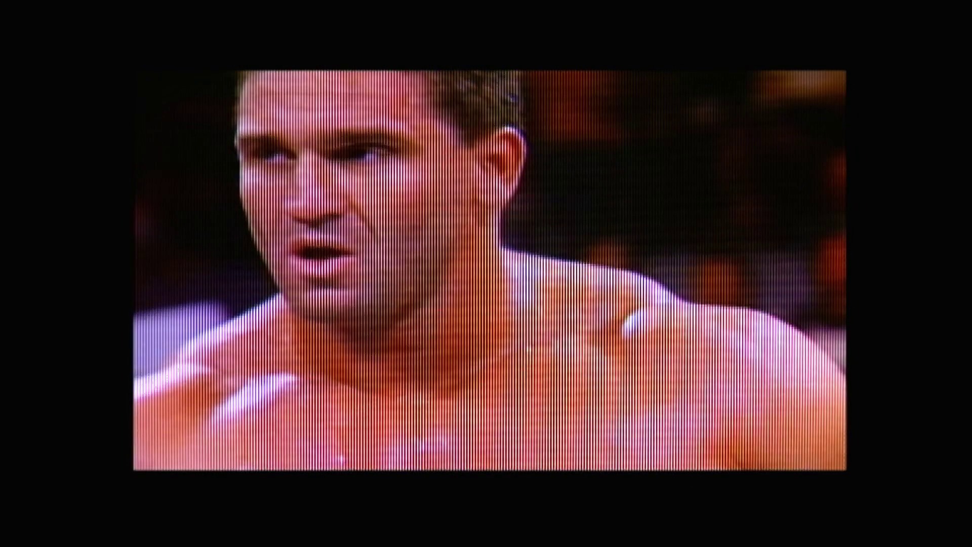 A frame from WWE Raw (November 23, 1998) at 0:50. A close-up shot of a wrestler's face displayed on a low-resolution TV screen with visible scan lines, characteristic of late 1990s broadcast quality. The wrestler appears shirtless against a blurred arena background in reddish and purple tones.