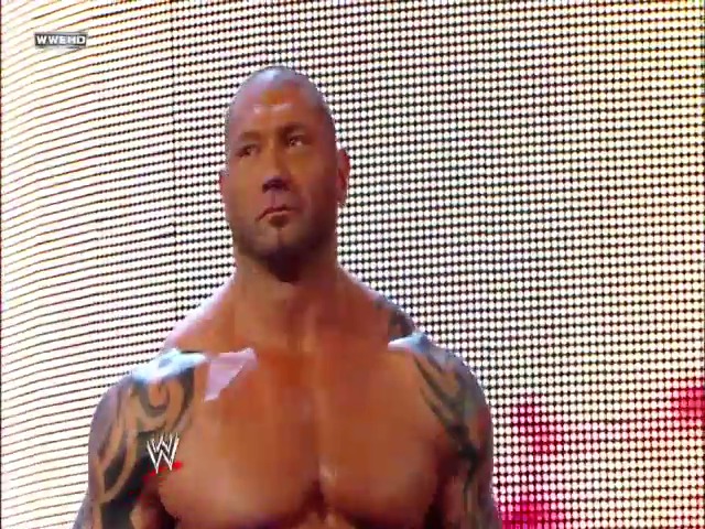 A frame from WWE SmackDown (January 1, 2010) at 1:11:21. A muscular male wrestler with tribal tattoos on his shoulders stands shirtless on a WWE broadcast, displayed on a large LED screen during SmackDown. The WWE logo is visible on his chest, and the pixelated screen backdrop fills the frame behind him.
