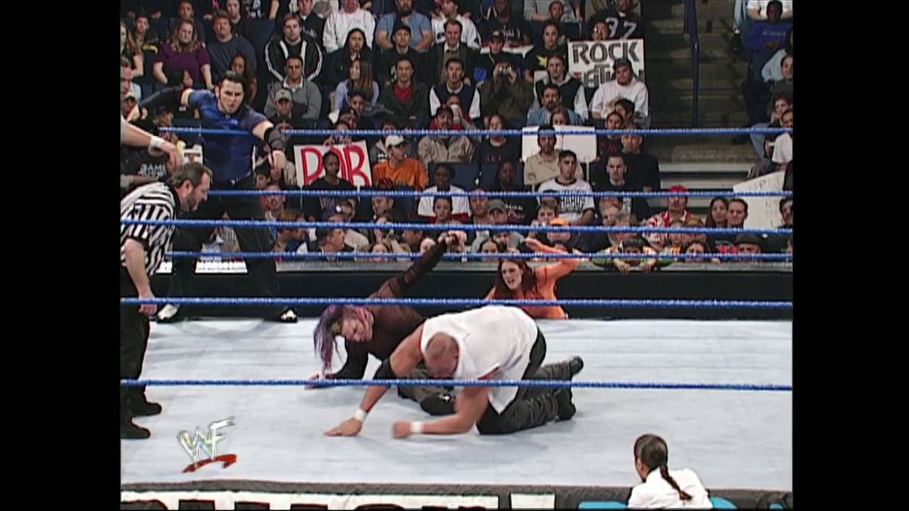 A frame from WWE SmackDown (January 11, 2001) at 23:26. A wrestler lies prone on the mat during a WWE SmackDown match, with an opponent nearby and a referee officiating. The blue-roped ring is surrounded by an enthusiastic crowd, with the WWF logo visible in the lower left corner.