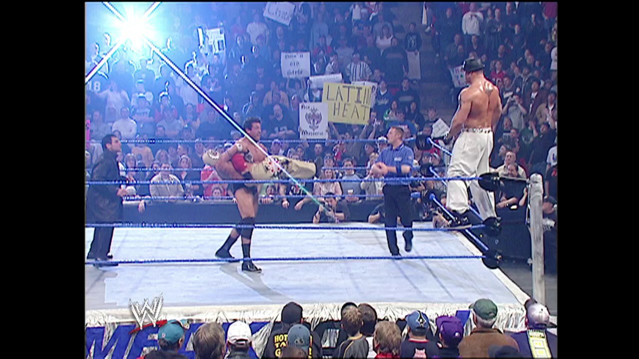A frame from WWE SmackDown (January 22, 2004) at 11:51. A WWE wrestler in white shorts stands on the ring apron while another performer executes a move in the blue-roped ring. The arena is packed with enthusiastic fans holding signs, with bright lighting and the WWE logo visible at the bottom of the screen.