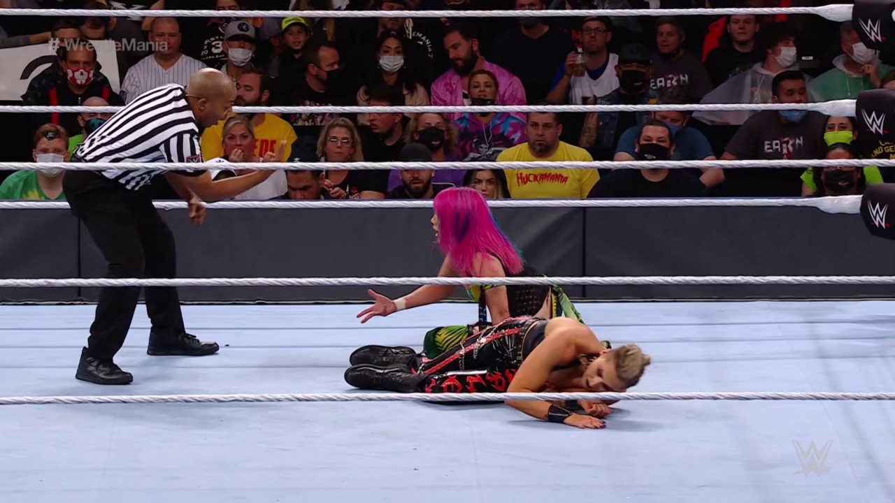 A frame from WWE WrestleMania 37 - Night 2 (2021) at 2:17:35. A WWE wrestler with bright pink hair lies on the canvas while another competitor stands nearby with an outstretched arm. A referee in a striped shirt officiates the match as a crowd of spectators watches from behind the ropes at WrestleMania.