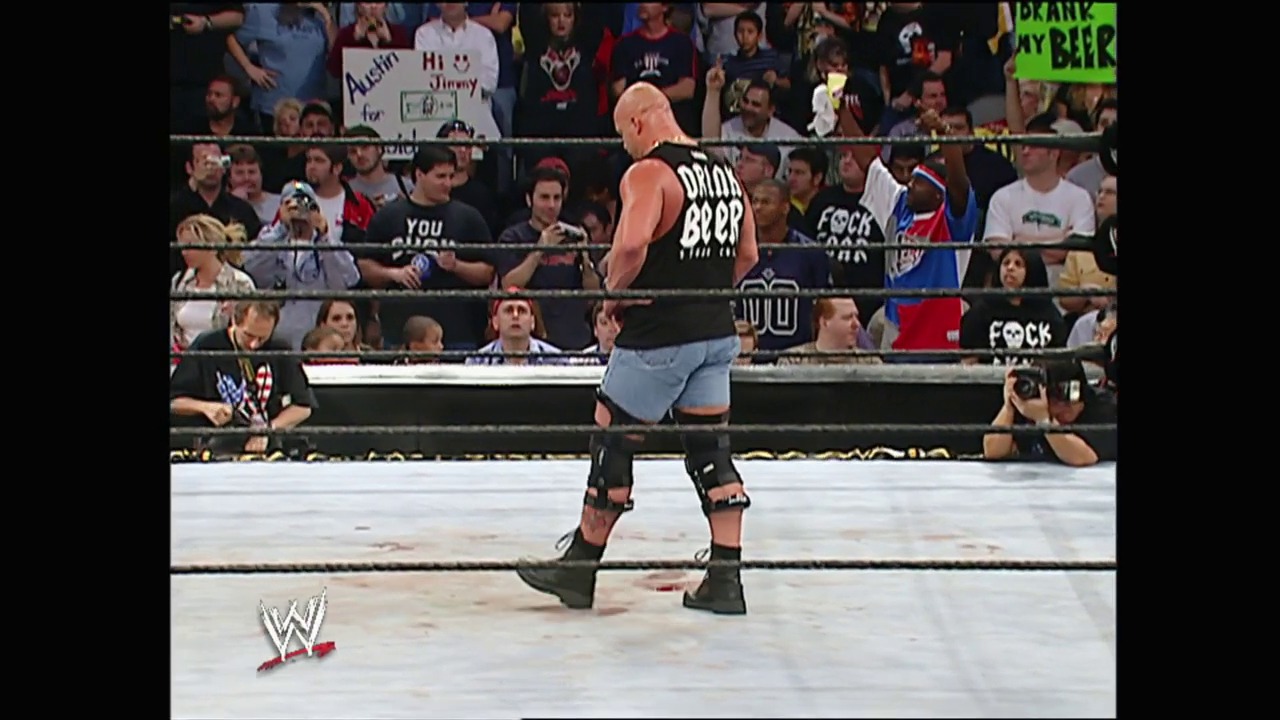 A frame from Survivor Series (2003) at 2:00:52. A bald wrestler stands in a WWE ring wearing a black sleeveless shirt and blue shorts, facing a crowd of spectators. The arena is filled with fans holding signs, with the WWE logo visible at the bottom of the frame.
