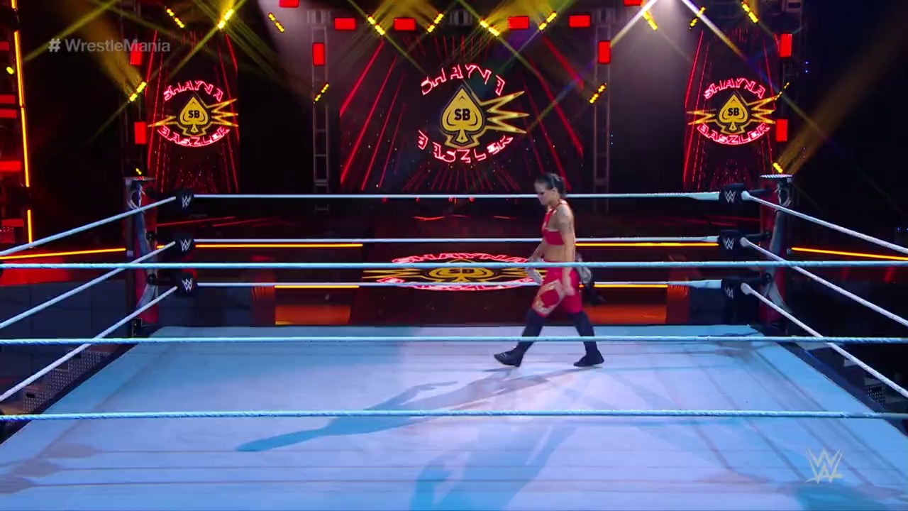 A frame from WrestleMania 36 (2020) at 53:41. A WWE wrestler in red and black attire stands in the ring during WrestleMania 36, with elaborate golden and red neon graphics displayed on the backdrop behind the blue-roped wrestling ring.