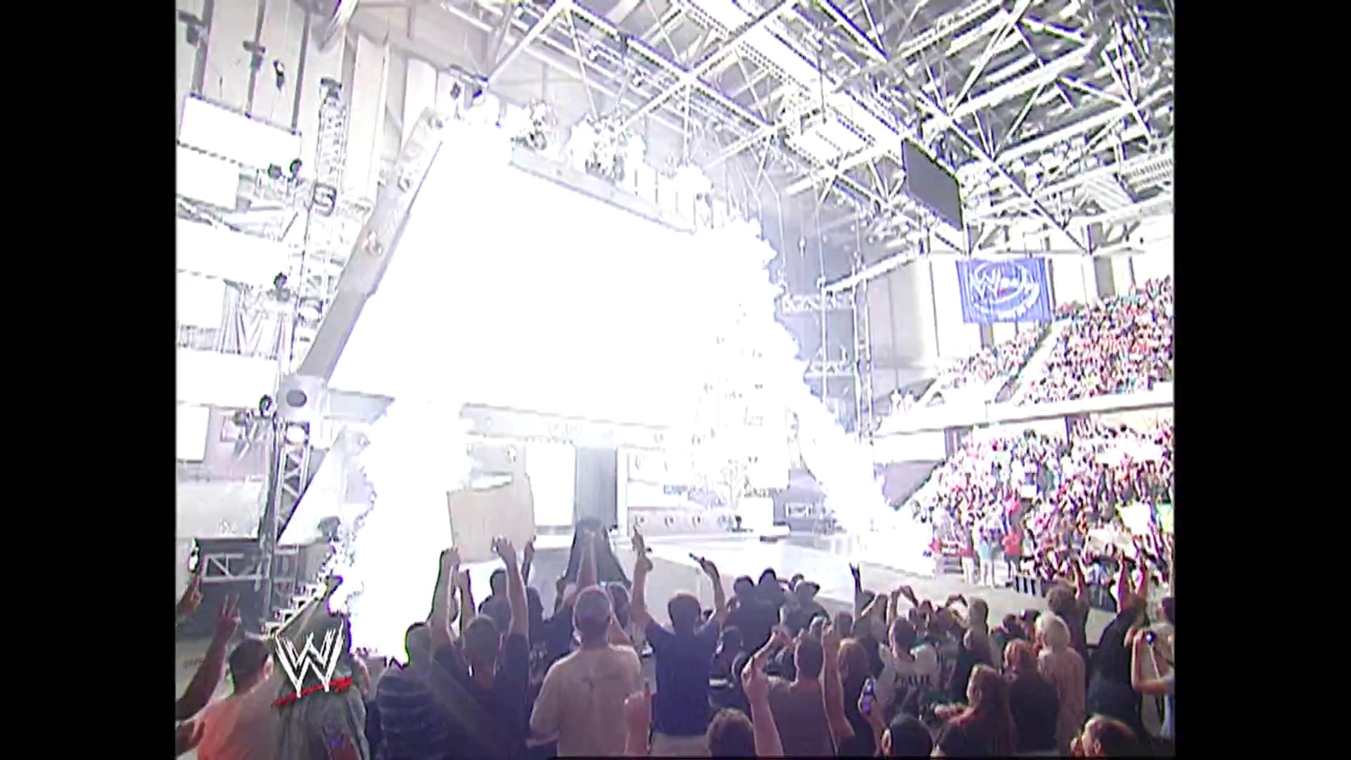 A frame from WWE Raw (October 2, 2006) at 0:52. A packed arena erupts with energy as thousands of fans fill the stands of a WWE wrestling event, with the illuminated ring visible in the center and the WWE logo prominent in the corner.