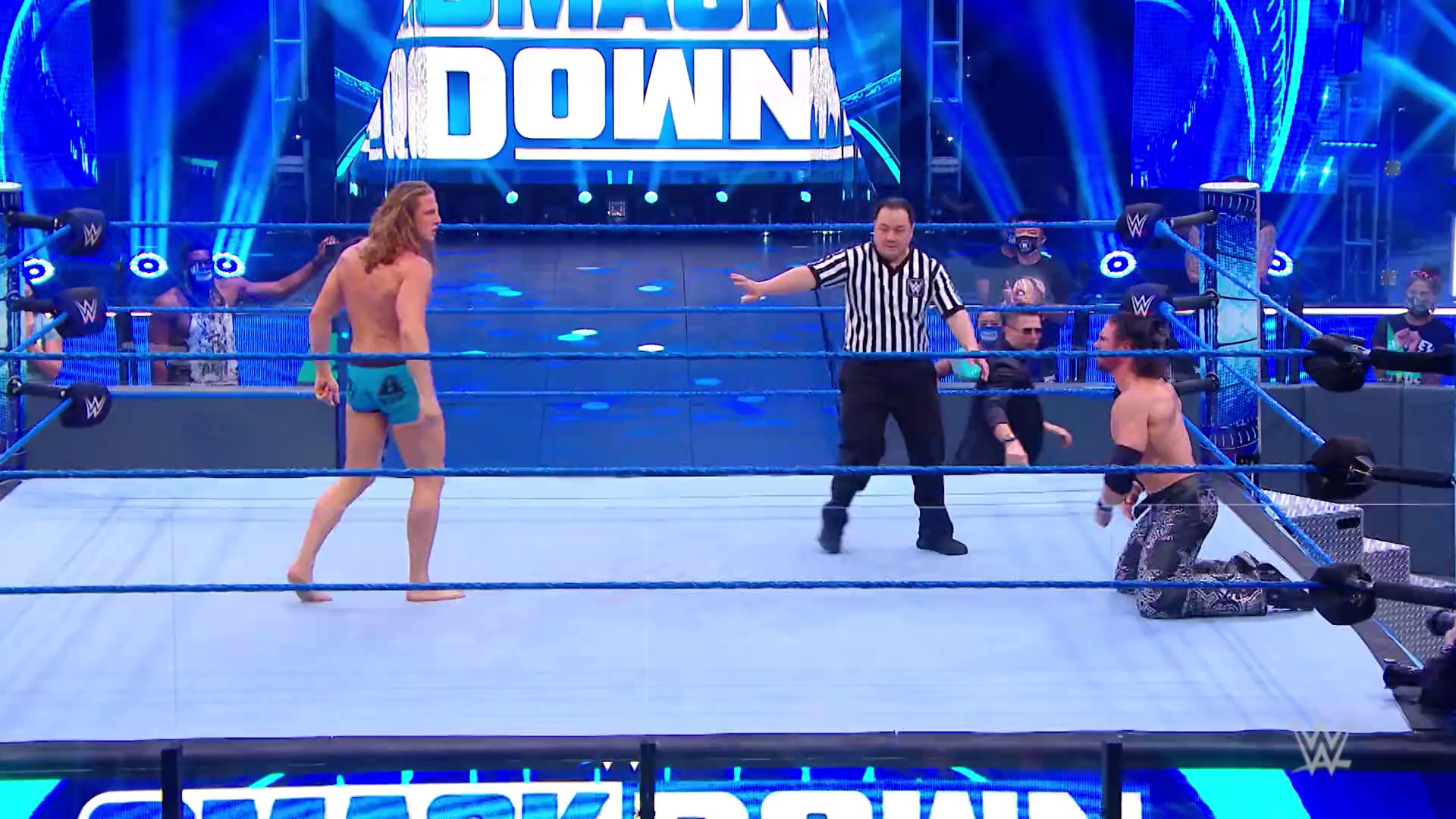A frame from WWE SmackDown (July 3, 2020) at 12:07. # WWE SmackDown Ring Scene

A WWE SmackDown match in progress features wrestlers in the blue-lit ring with a referee in striped shirt officiating. The illuminated "SMACK DOWN" graphics are visible above the ring, with audience members seated behind the barriers.