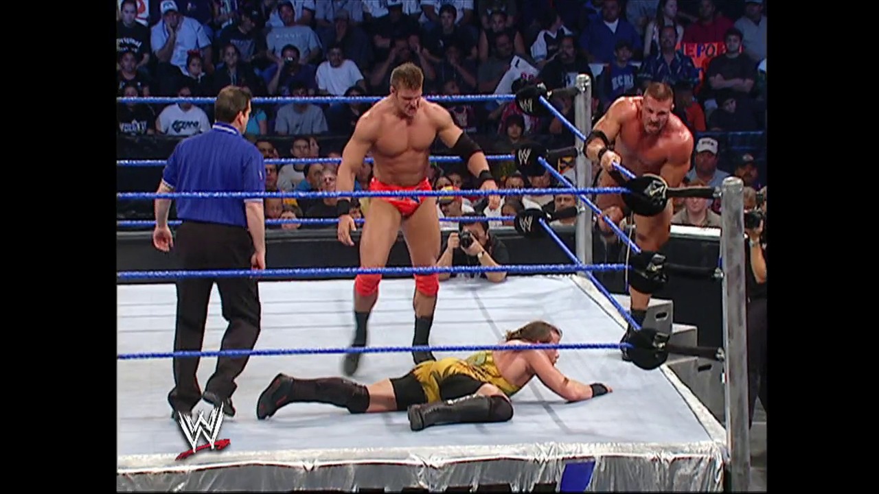 A frame from WWE SmackDown (November 11, 2004) at 49:23. A WWE wrestler in yellow and black lies prone on the white mat while two opponents in red and orange trunks stand nearby, with a referee in blue officiating. The blue-roped ring is surrounded by an engaged crowd under bright arena lighting.