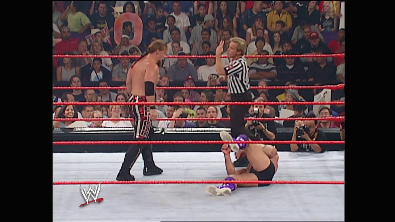 A frame from WWE Unforgiven (2002) at 23:47. A WWE wrestling match in progress inside a red-roped ring with a shirtless wrestler standing while another lies on the mat. A referee in striped shirt officiates as a packed crowd watches from the stands.