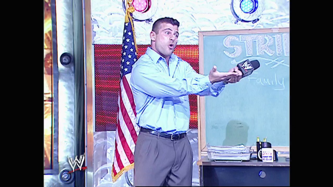 A frame from WWE Raw (December 12, 2005) at 1:04:37. A man in a light blue shirt and gray pants stands in an office setting with an American flag behind him, holding what appears to be a WWE championship belt. The WWE logo is visible in the bottom left corner of the frame.