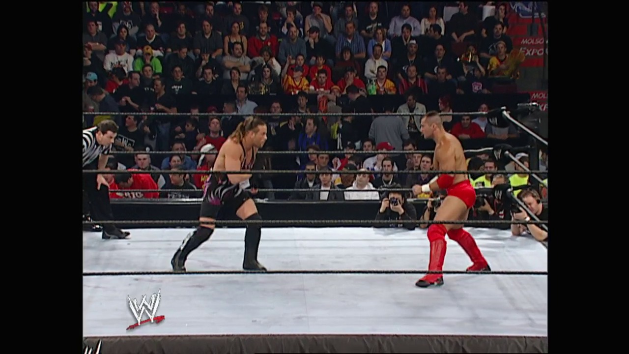 A frame from WWE No Way Out (2003) at 22:57. Two wrestlers face off in a WWE ring during No Way Out 2003, with one in black attire and the other in red pants. The crowd fills the arena stands behind the black ropes as the referee watches the match unfold.