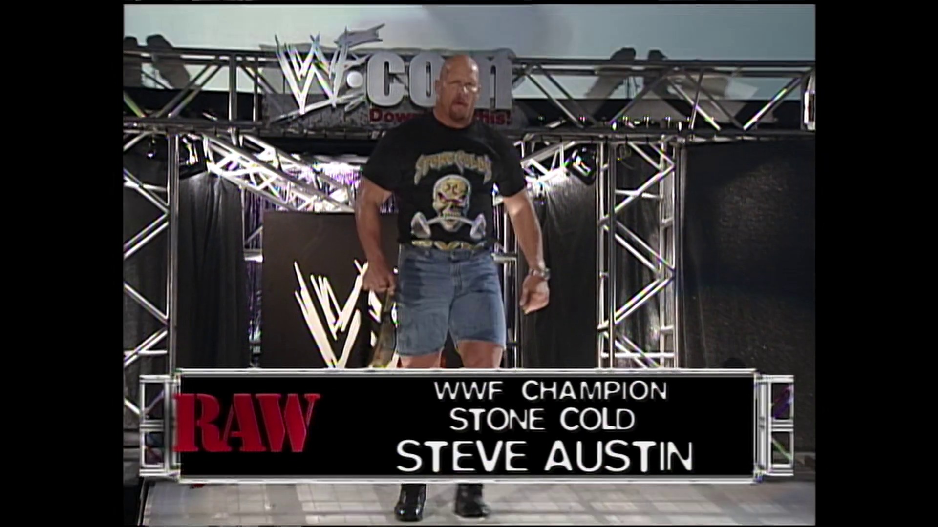 A frame from WWE Raw (July 12, 1999) at 1:47. WWE Raw entrance scene featuring Stone Cold Steve Austin, the WWF Champion, walking to the ring in a black skull graphic t-shirt and denim shorts. The stage displays WWF branding with steel framework and lighting, with the RAW logo and character information visible at the bottom of the screen.