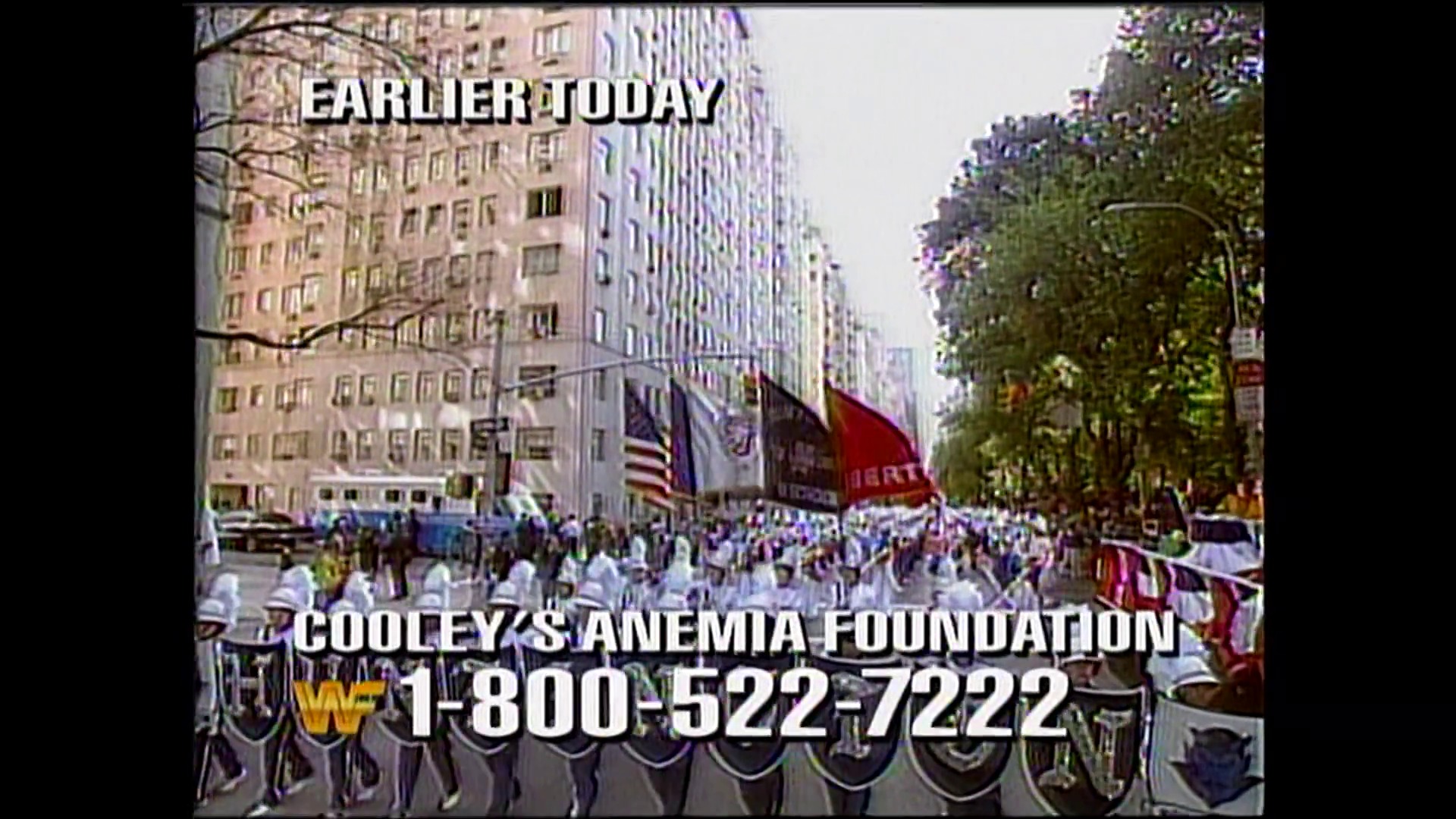 A frame from WWE Raw (October 10, 1994) at 21:29. A large parade or public event fills a city street lined with tall buildings, featuring crowds of people in white uniforms and colorful flags. On-screen graphics display "EARLIER TODAY" and charity information for Cooley's Anemia Foundation with a phone number.