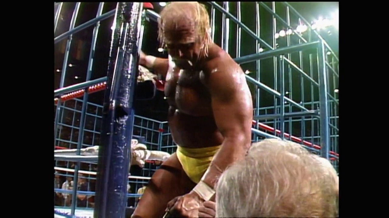 A frame from WrestleMania 2 (1986) at 3:01:11. # WrestleMania 2 (1986) - Frame Description

A muscular wrestler with blonde hair, wearing yellow trunks, grapples with an opponent against the blue steel cage during an intense wrestling match, with arena lighting and crowd visible in the background.