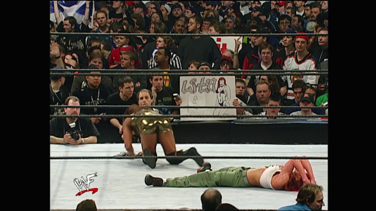 A frame from WrestleMania X8 (2002) at 3:10:27. A wrestler in gold attire kneels in the ring while another competitor lies on the mat in green and red clothing. The crowd fills the stands behind the ropes, with the WWF logo visible in the corner.