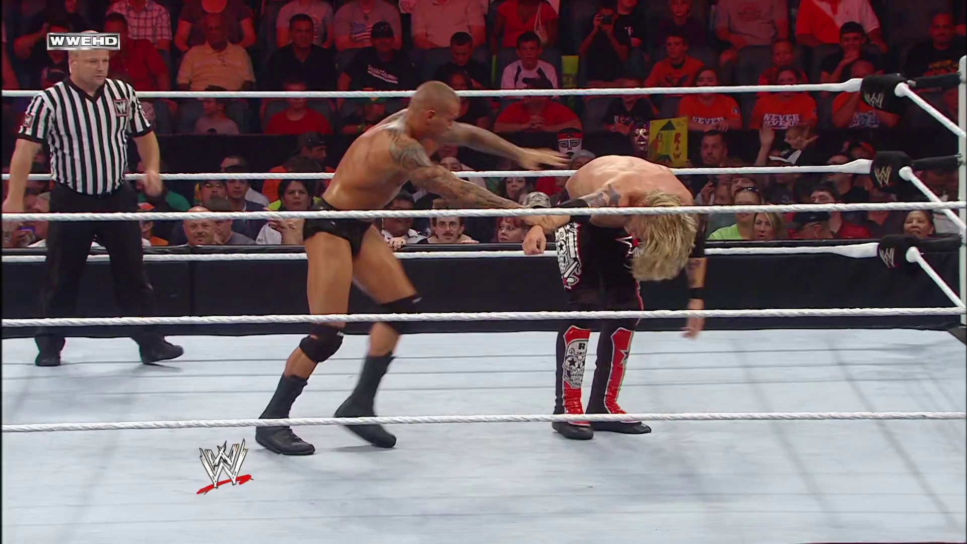 A frame from WWE Raw (July 19, 2010) at 13:38. Two wrestlers face off in a WWE Raw match inside the white-roped ring, with a referee in striped shirt overseeing the action. A packed crowd fills the arena in the background as the WWE logo appears in the bottom left corner.