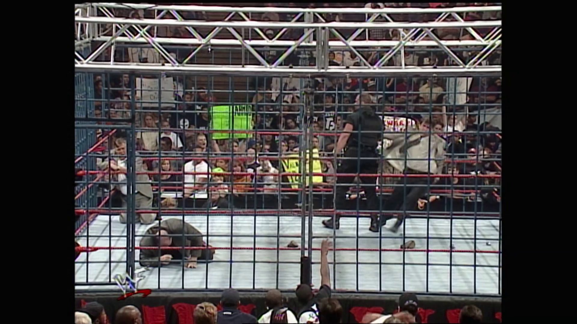A frame from WWE Raw (November 2, 1998) at 1:30:41. A wrestling match unfolds inside a steel cage with spectators packed in the stands above and around the ring. Two wrestlers are visible on the mat while officials and audience members watch intently from behind the cage barriers.