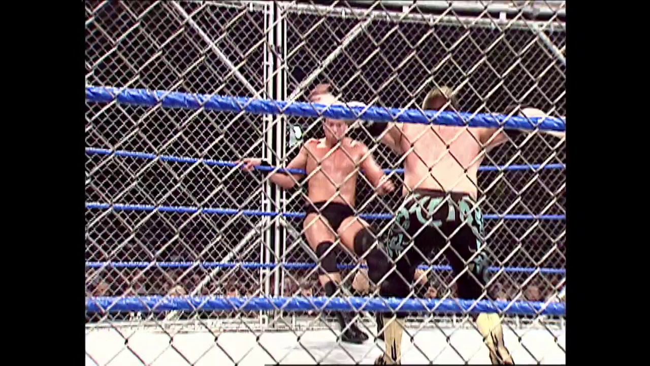 A frame from WWE SmackDown (July 22, 2004) at 0:51. A shirtless wrestler stands inside a steel cage match setup with blue ring ropes visible. The chain-link fence structure dominates the frame, creating a barrier between the performer and the audience during this WWE SmackDown event.