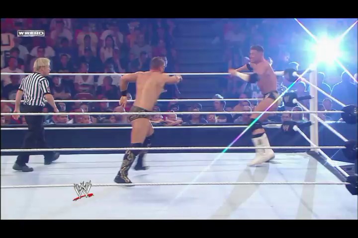 A frame from WWE Raw (July 4, 2011) at 1:06:27. Two shirtless wrestlers grapple in a WWE ring during an intense match, with a referee in striped shirt nearby and a crowd visible in the background. Bright arena lighting illuminates the white ring as one competitor extends his arms while facing his opponent.