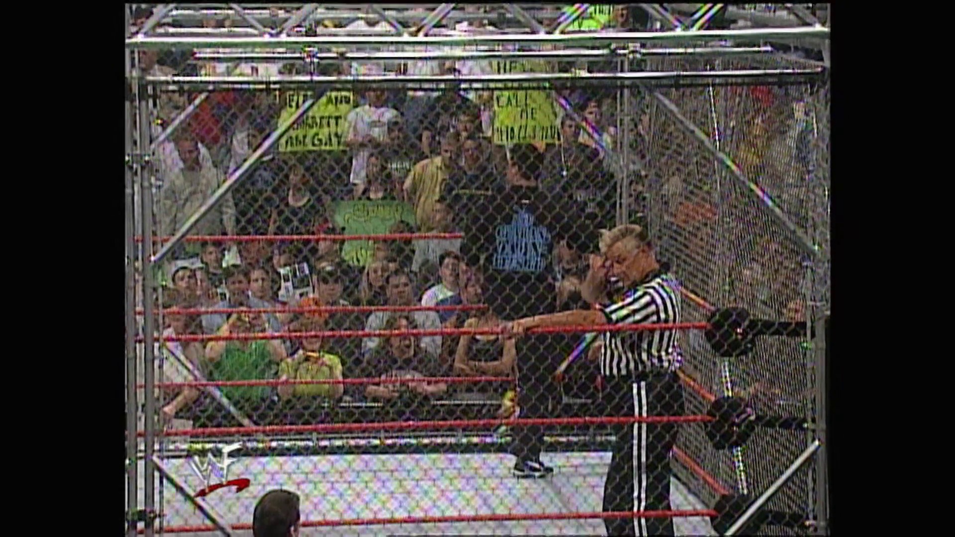 A frame from WWE Raw (May 1, 2000) at 1:22:14. A referee in a striped shirt stands in a steel cage wrestling ring, gesturing toward the crowd. The cage is surrounded by spectators holding signs, with the red ropes and mesh fencing clearly visible in this WWE Raw broadcast.