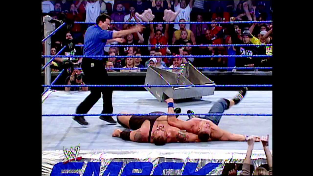 A frame from WWE SmackDown (November 20, 2003) at 1:25:33. A WWE wrestler lies prone on the blue mat while another wrestler in blue clothing stands nearby, with a steel trash can visible in the ring. The crowd fills the arena stands in the background, and the WWE logo is visible at the bottom of the screen.