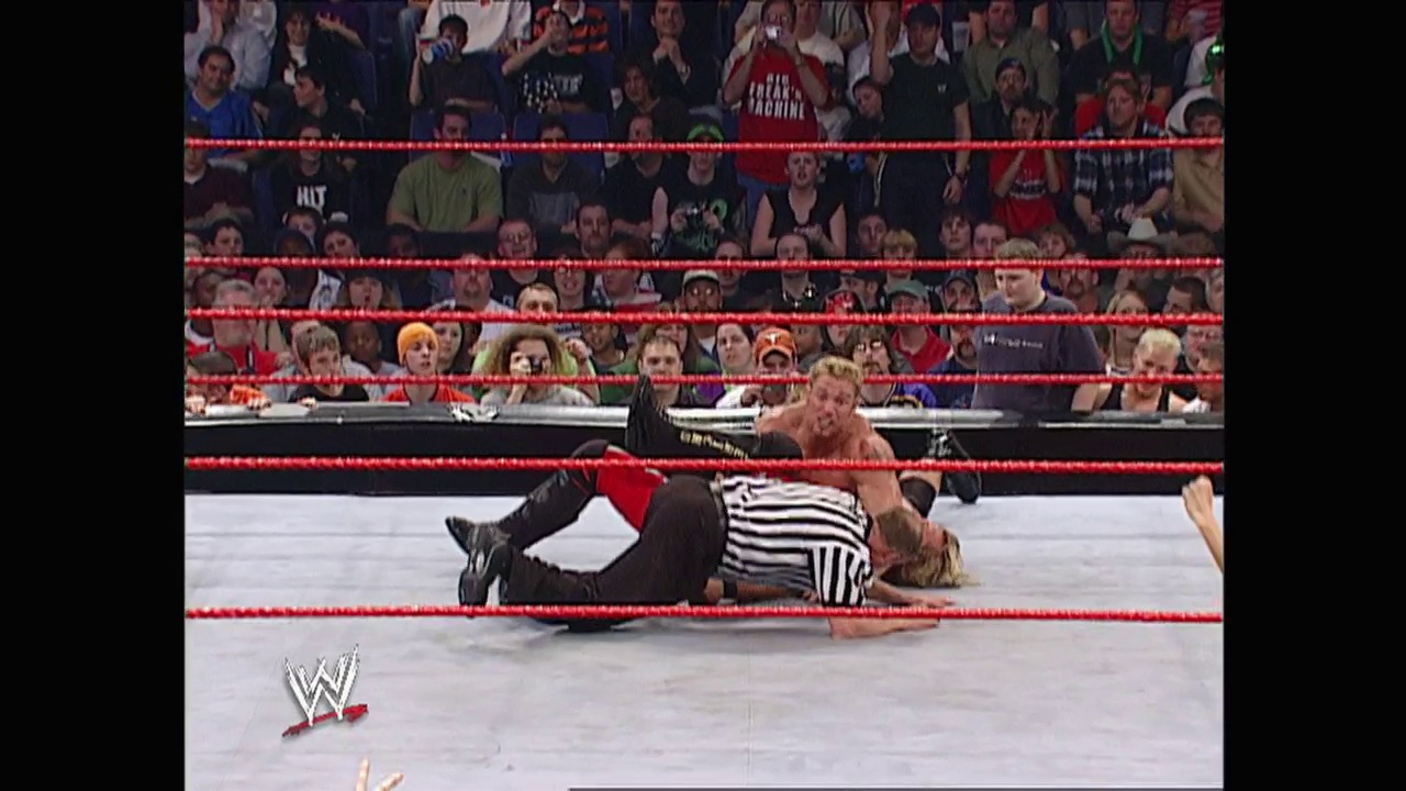 A frame from WWE Raw (March 17, 2003) at 1:06:51. A wrestler lies on the mat in a WWE Raw ring surrounded by red ropes, while a referee in a striped shirt stands nearby. The crowd fills the arena seats in the background, with the WWE logo visible in the corner.