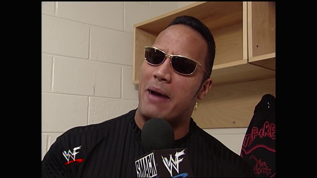 A frame from WWE SmackDown (September 30, 1999) at 17:52. A wrestler wearing dark sunglasses and black WWF attire speaks into a microphone backstage. The scene takes place in a corridor with white brick walls and wooden shelving visible in the background.