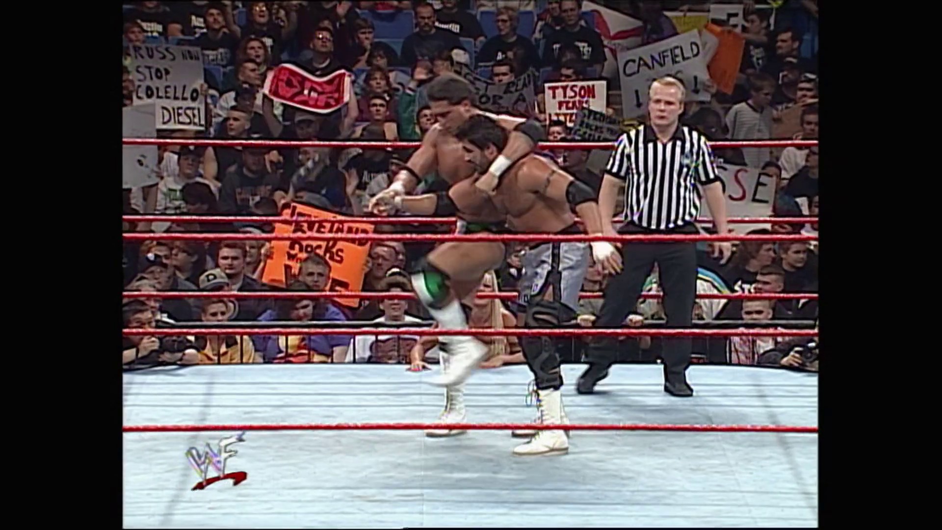 A frame from WWE Raw (March 2, 1998) at 26:38. A WWE Raw wrestling match in progress, with two wrestlers in the ring—one performing a move on the other while a referee in black and white stripes watches. The crowd fills the arena behind the red-roped ring, holding signs including references to Tyson and Canfield.