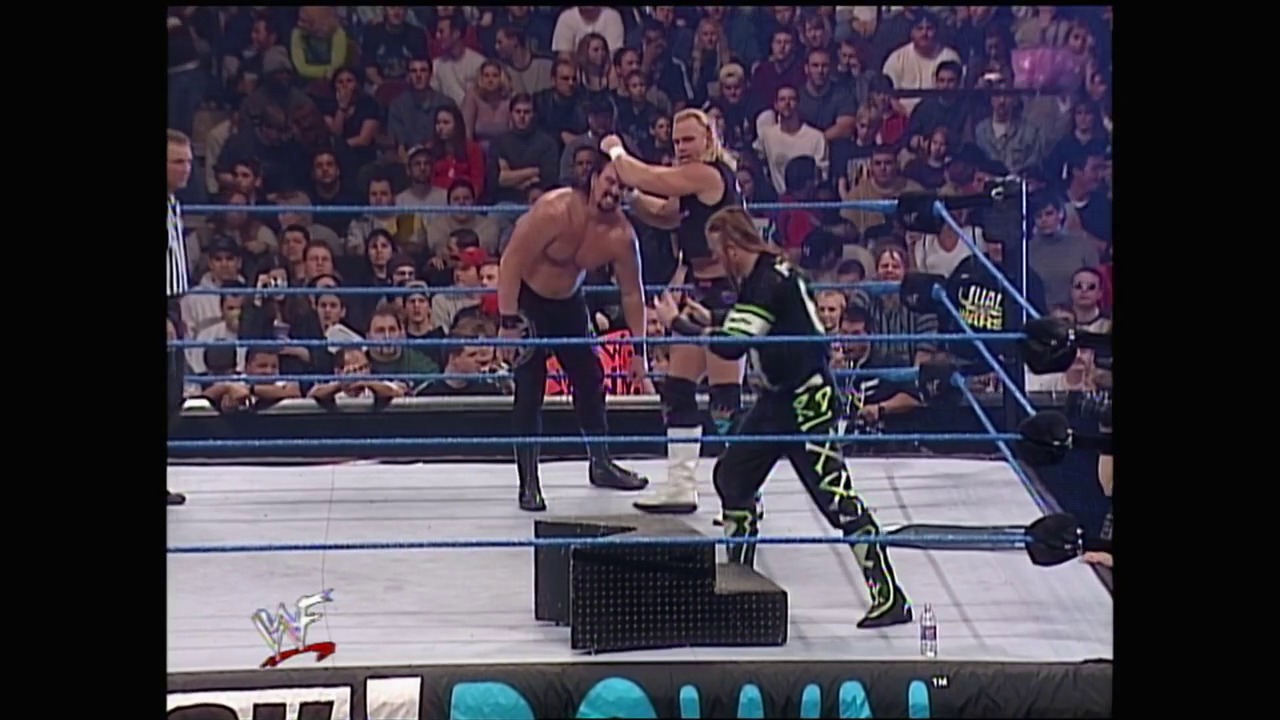 A frame from WWE SmackDown (January 13, 2000) at 27:16. Two wrestlers face off in a WWE ring during an intense match, with one shirtless competitor in dark pants and another in black attire with neon green accents. A full crowd watches from the stands as the referee oversees the action on the blue-roped ring.