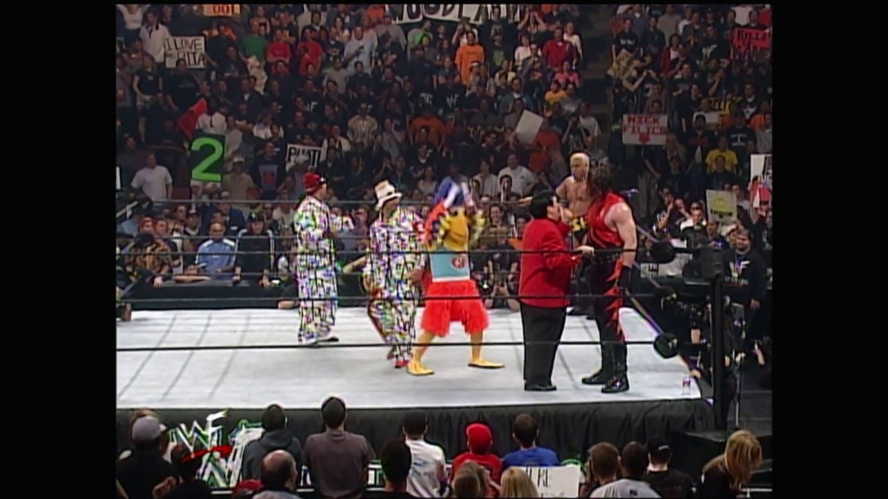 A frame from WrestleMania 2000 (2000) at 2:21:23. A multi-team wrestling match is underway in the WWF ring, with wrestlers in colorful attire (checkered patterns, red gear) facing off while a packed crowd watches from the stands. The arena displays WWF branding and signage throughout the venue.