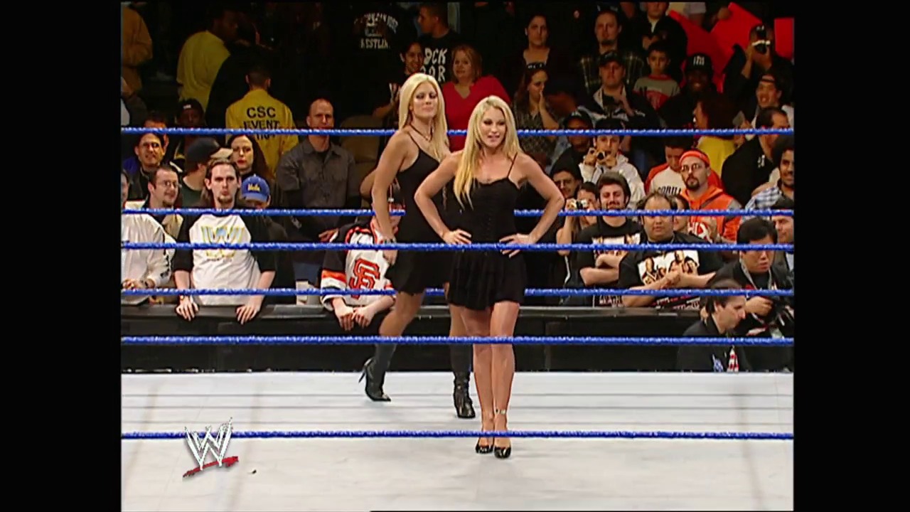 A frame from WWE No Way Out (2004) at 0:49. Two blonde female wrestlers stand in a WWE ring during No Way Out 2004, with one posing confidently while the other stands behind. The crowd fills the arena stands surrounding the blue-roped ring, with the WWE logo visible in the corner.