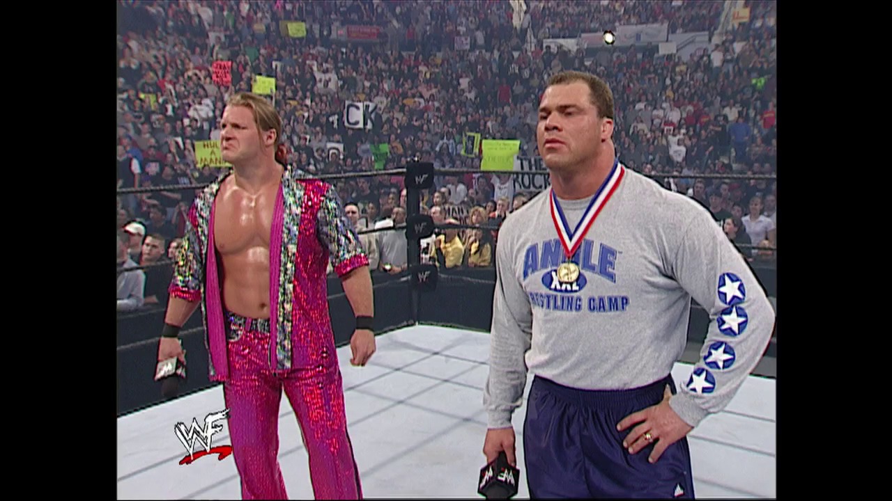 A frame from WWE Raw (April 4, 2002) at 12:45. Two wrestlers stand in the WWE ring during Raw, with one wearing a hot pink sequined jacket and the other in an "Angle Wrestling Camp" gray sweatshirt. A packed crowd fills the arena in the background behind the white-roped ring.