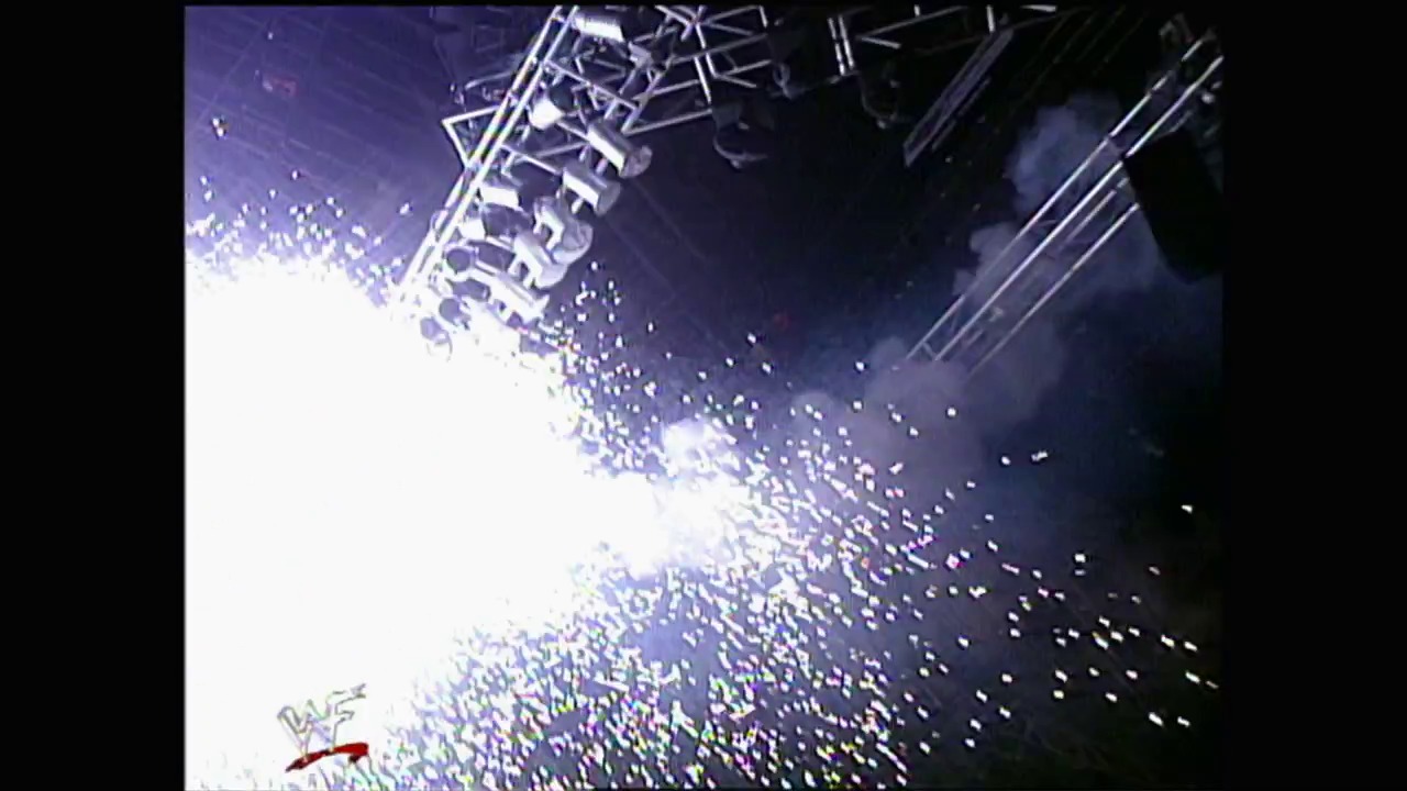A frame from WWE SmackDown (September 16, 1999) at 1:03. A massive crowd fills an arena during a WWE event, with pyrotechnics exploding above the ring and stage structure. The bright flash of fireworks illuminates the sea of spectators in the dimly lit venue.