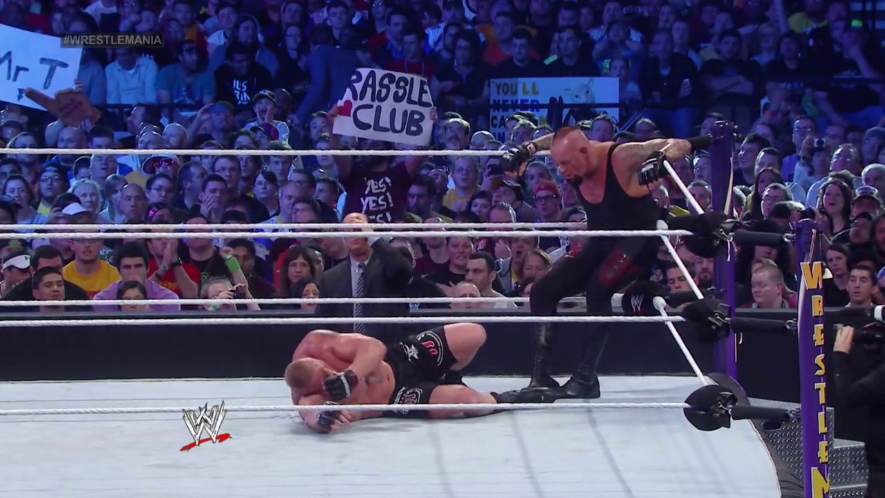 A frame from WrestleMania XXX (2014) at 2:46:51. A WWE wrestler lies on the mat while another stands on the ring apron during WrestleMania XXX. The crowd fills the arena with signs visible, including a "Rassle Club" banner, while the WWE logo appears at the bottom of the frame.