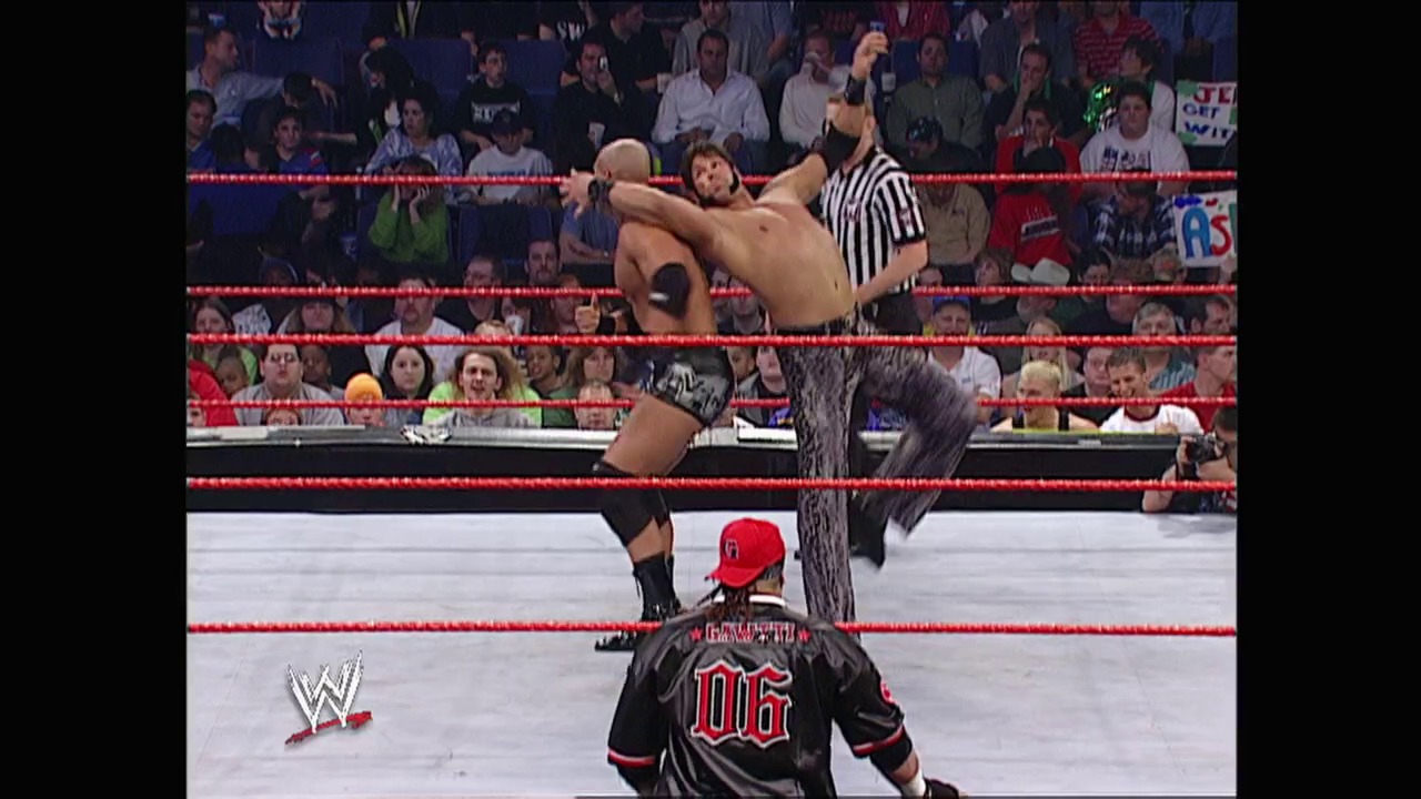 A frame from WWE Raw (March 17, 2003) at 27:29. Two wrestlers are engaged in mid-action inside a WWE ring bordered by red ropes, with one performer executing an aerial move while another waits below. A referee in black and white stripes oversees the match as a packed crowd watches from the stands.