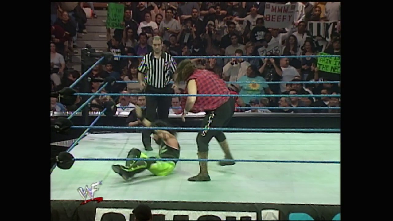 A frame from WWE SmackDown (February 17, 2000) at 47:21. A WWE wrestler in a bright neon green outfit lies on the mat while an opponent in a red/maroon shirt stands over them. A referee in stripes oversees the match inside the blue-roped ring, with an enthusiastic crowd visible in the background.