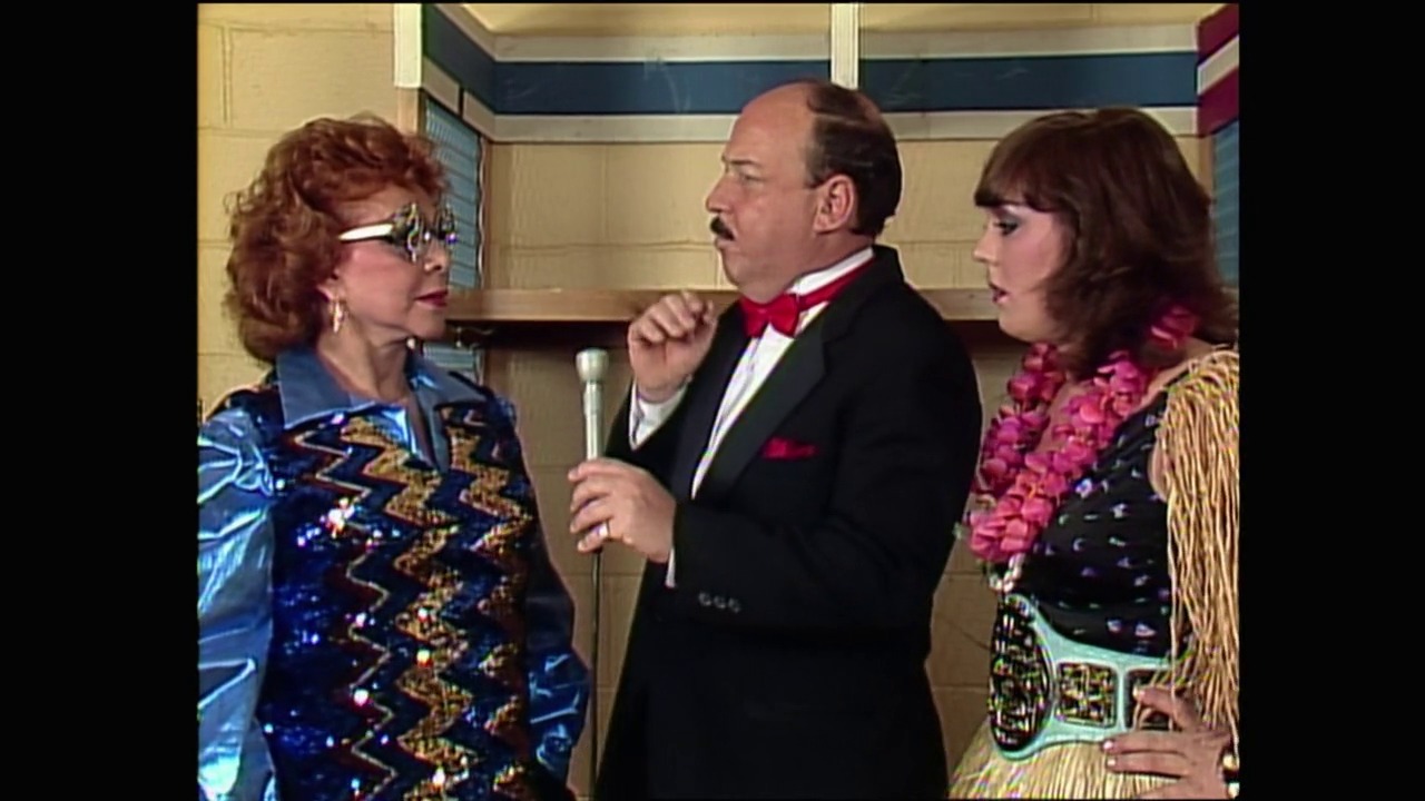 A frame from WrestleMania I (1985) at 1:28:38. A man in a tuxedo with a microphone stands between two women in colorful, sequined outfits in what appears to be a backstage interview setting. The scene captures a moment from a professional wrestling event with typical 1980s styling and décor.