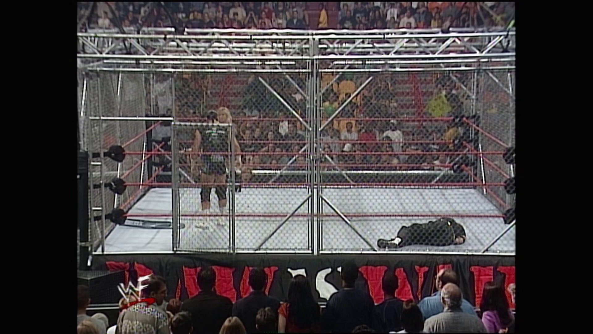 A frame from WWE Raw (January 3, 2000) at 1:09:09. A wrestler lies on the mat inside a steel cage during a WWE Raw match, while another competitor stands nearby. The packed arena crowd watches from the stands as the WWF logo is visible on the barrier below.