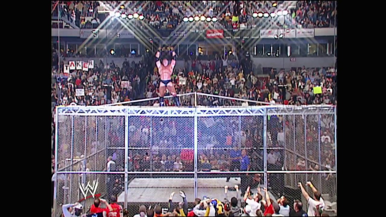 A frame from WWE No Mercy (2002) at 2:45:57. A wrestler stands triumphantly atop the steel cage structure during a WWE match, with an energetic crowd visible in the background stands and the illuminated arena setting the dramatic scene.