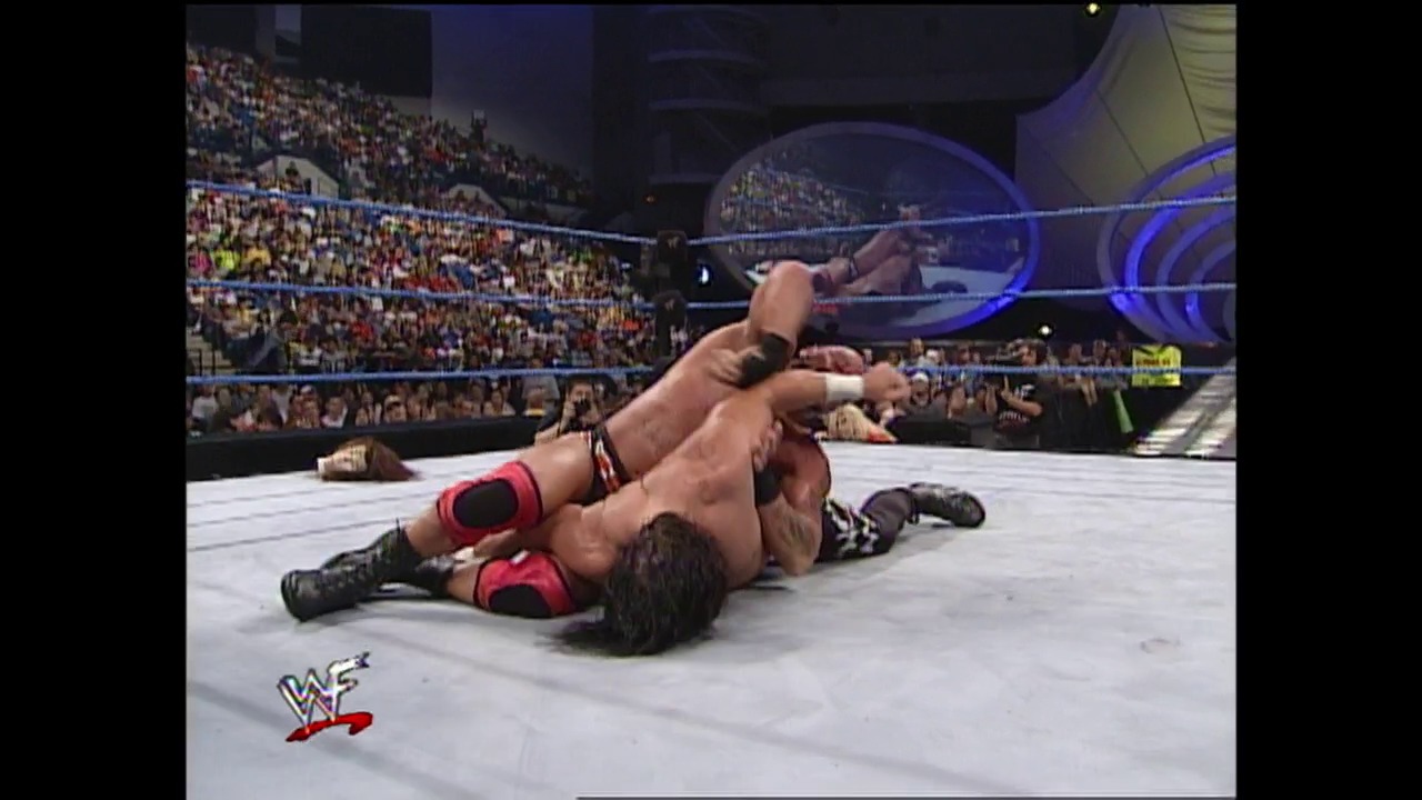 A frame from WWE SmackDown (August 31, 2000) at 38:04. A wrestler in red and black attire executes a submission move on their opponent on the white canvas, while a packed arena crowd watches from the stands. The WWE SmackDown logo is visible in the lower left corner of the broadcast.