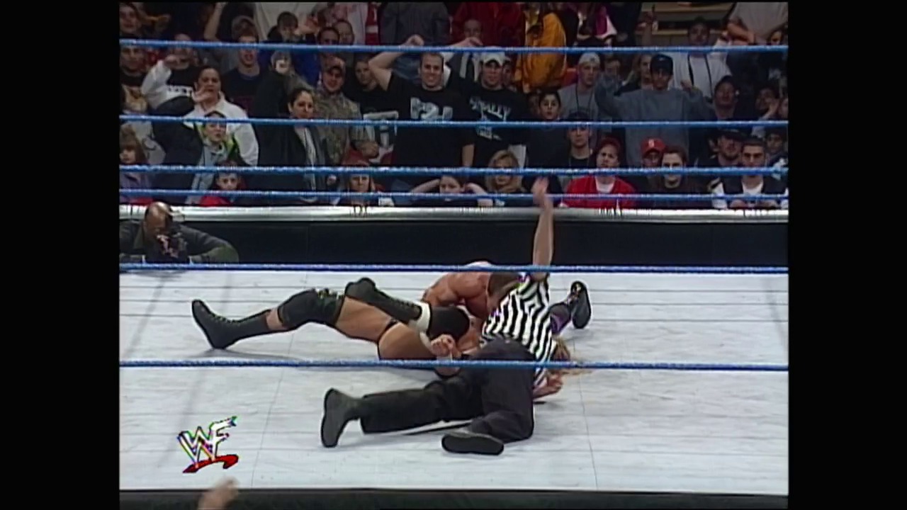 A frame from WWE SmackDown (February 3, 2000) at 1:34:11. A wrestler lies pinned on the mat while a referee in a striped shirt counts, with a large crowd visible in the bleachers behind the blue-roped ring. The WWE logo appears in the lower left corner of the broadcast.
