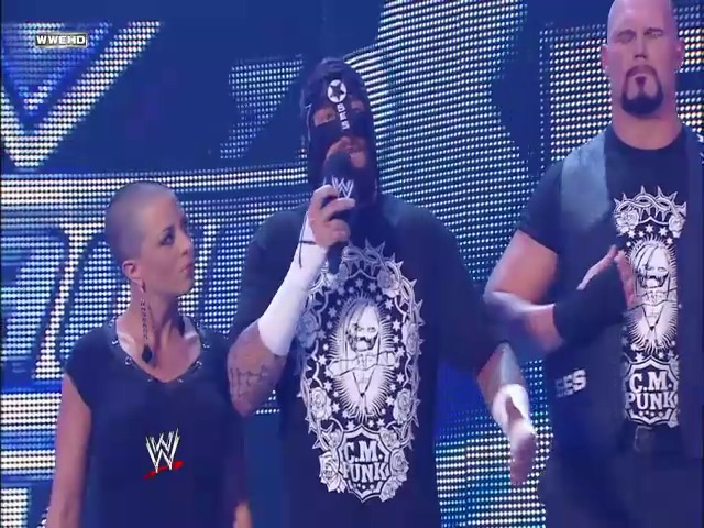 A frame from WWE SmackDown (June 11, 2010) at 12:18. Three WWE wrestlers stand in the ring wearing matching skull graphic t-shirts, with one holding a microphone. A large blue LED screen backdrop displays the WWE logo and graphics behind them.